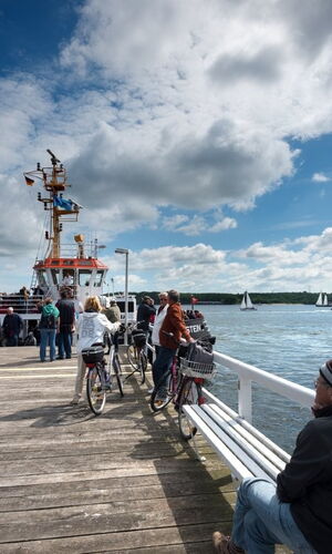 Menschen auf einem Holzsteg neben einem Hafen mit mehreren Segelbooten und einem großen Schiff unter bewölktem Himmel